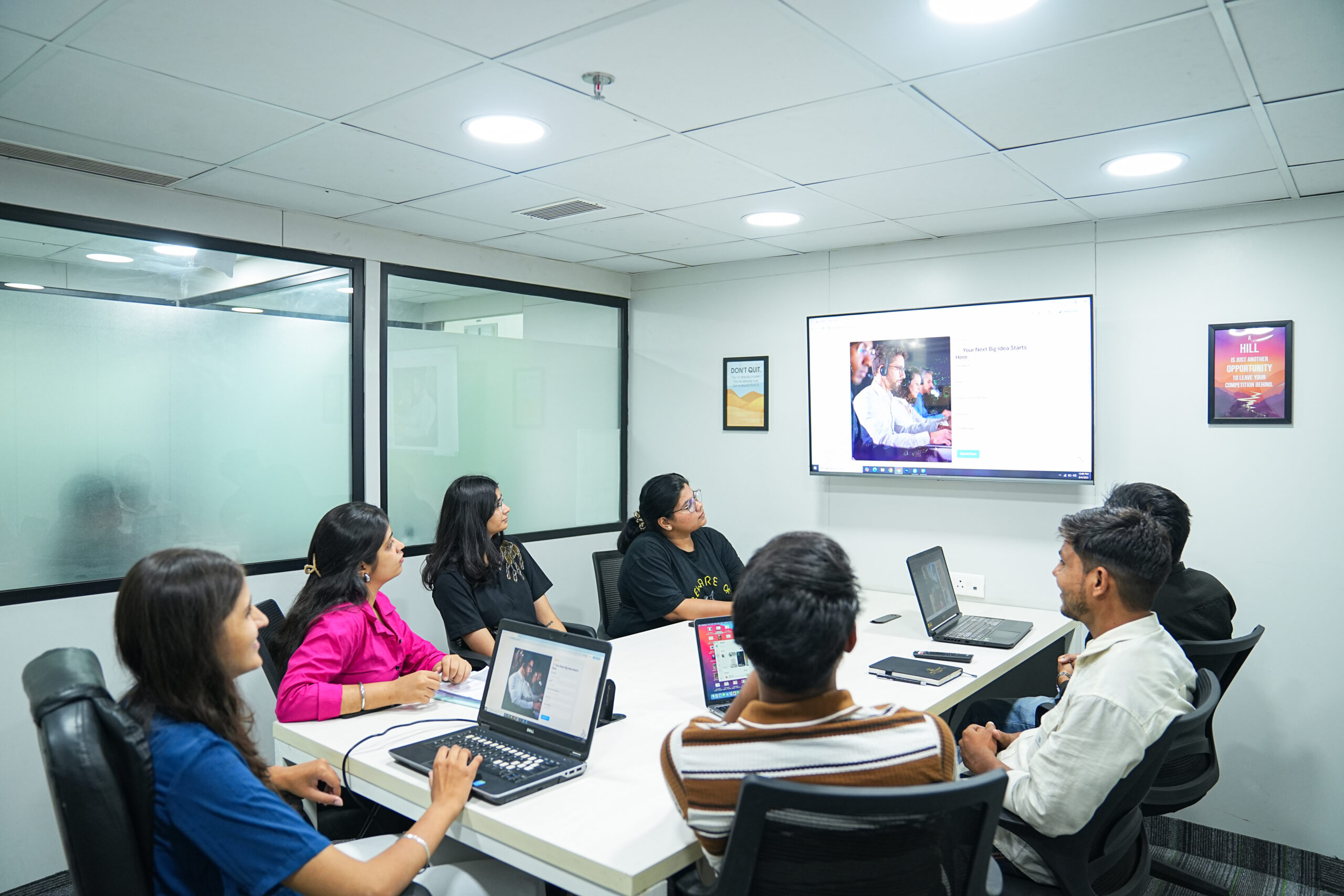 Six people sit around a conference table with laptops, attentively watching a presentation by a leading social media marketing agency in India in a modern office meeting room.